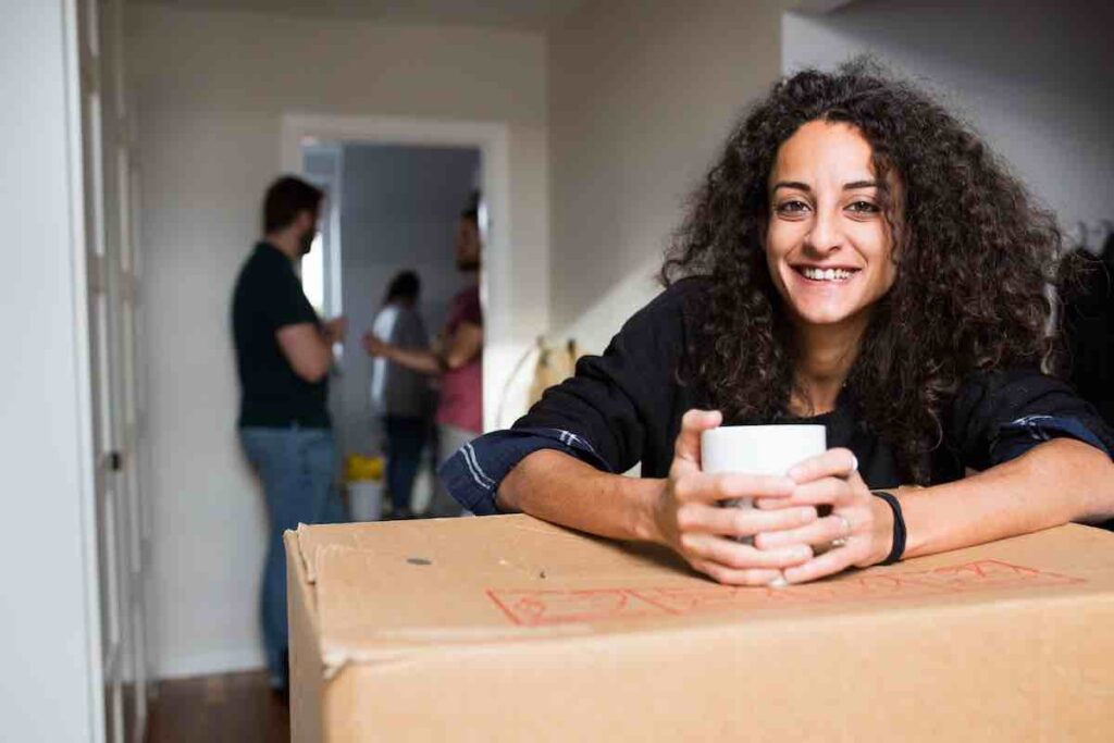 Happy woman leaning on a box, relaxing after workingHappy woman leaning on a box, relaxing after working through her moving checklist PDF.through her moving checklist PDF.